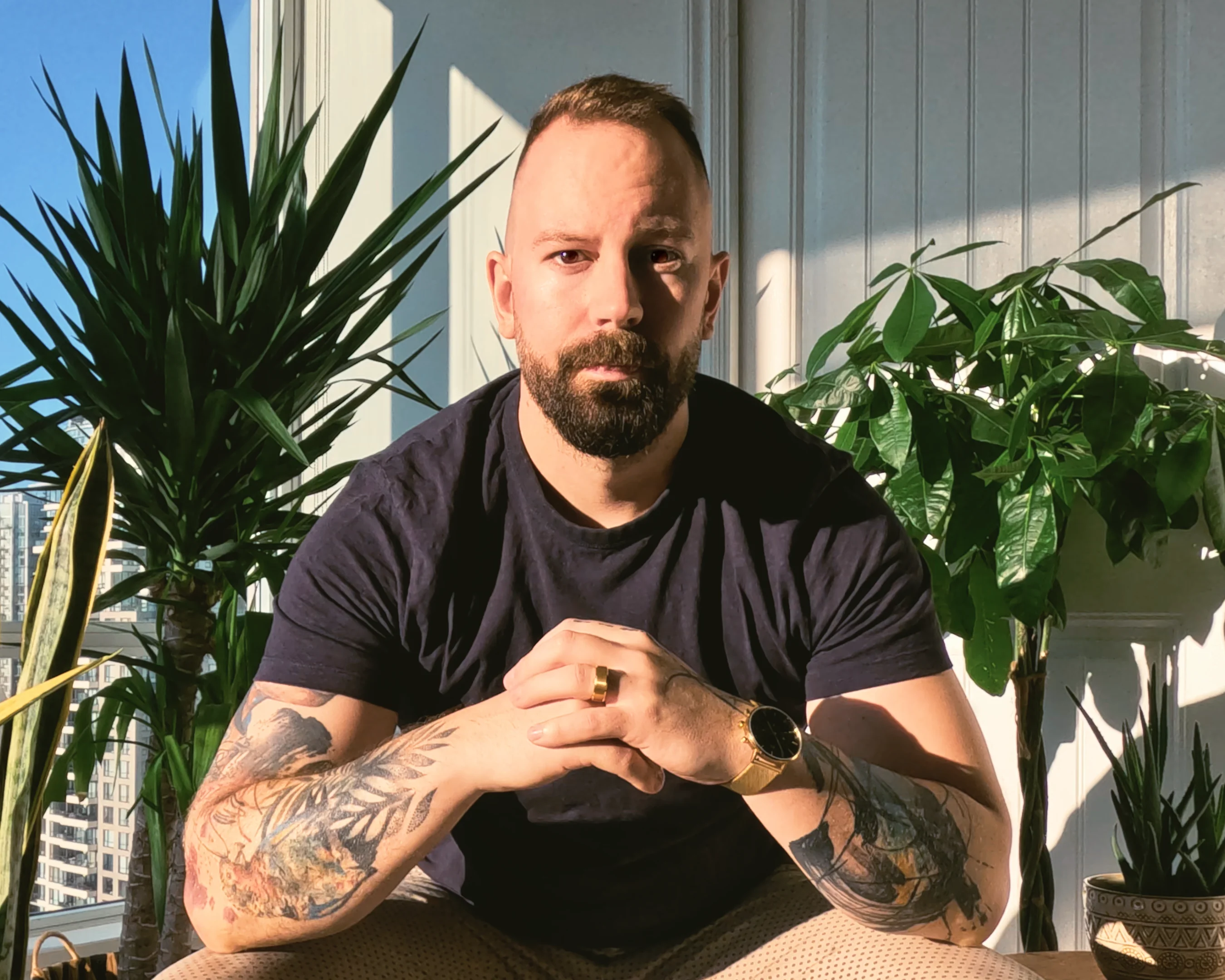 A white man in his mid-thirties sitting in front of tropical trees and plants. The man has a beard, a short haircut, and is wearing a dark blue t-shirt. His arms are folded in font of him and depict tattoos of stylized brushwork and a colorful parrot. The man is wearing a golden wedding ring and a matching golden watch.