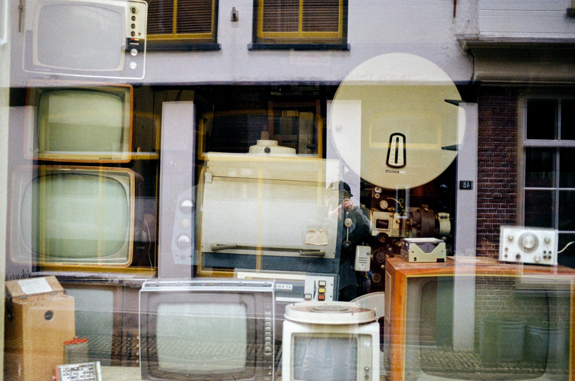 Self-portrait in a shop mirror showing vintage TVs and a film projector inside.